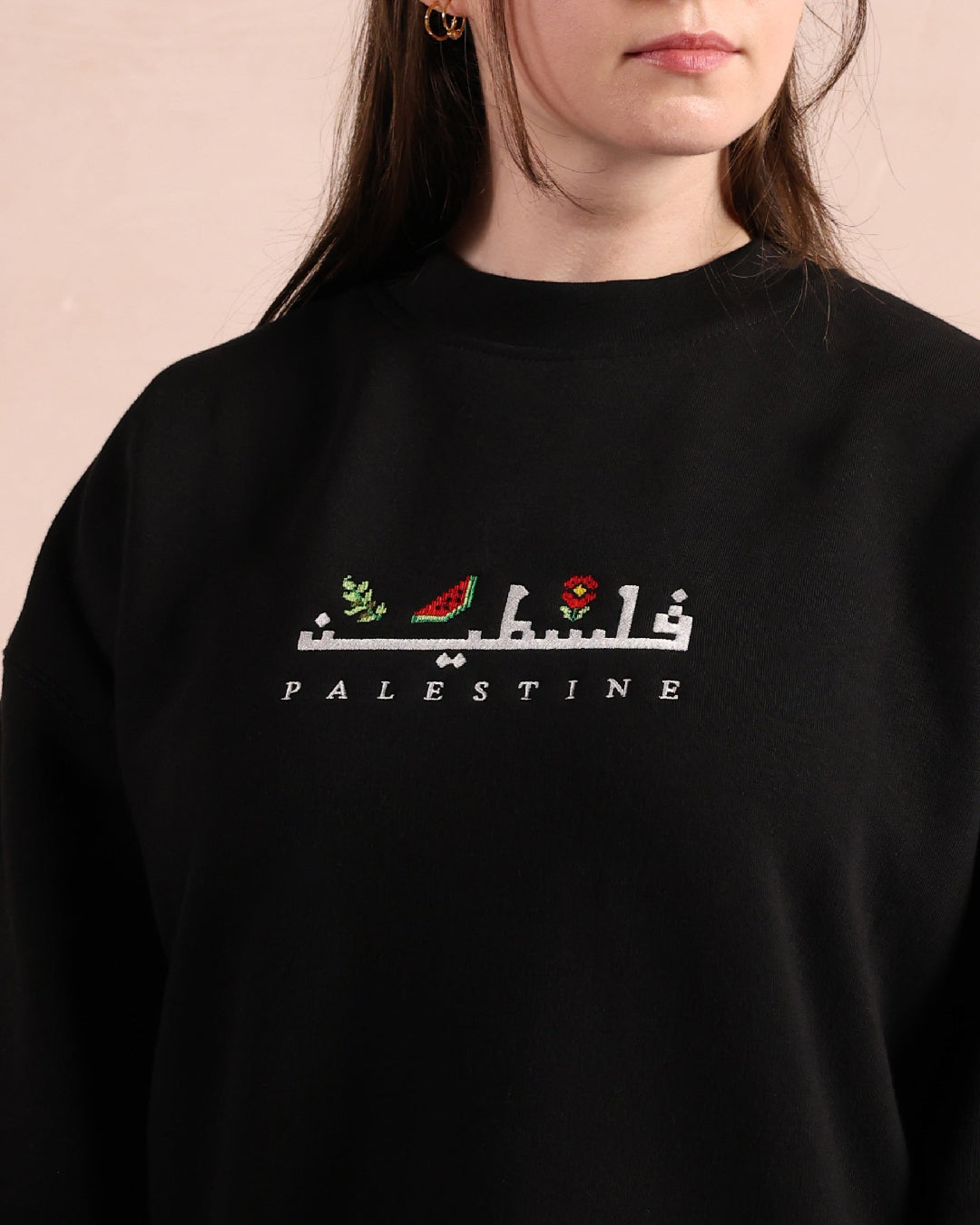 a closeup of a woman wearing a black sweatshirt embroidered with the word palestine in english and arabic in white alongside an olive branch, red poppy and watermelon