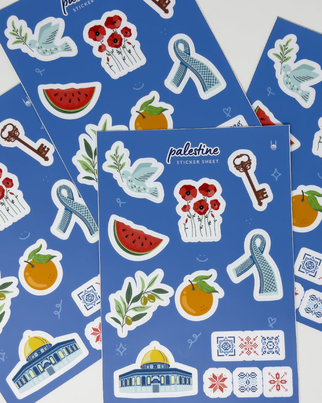 A collection of blue sticker sheets layered on top of each other, with designs inspired by Palestine, including a dove holding an olive branch, poppies, a key, keffiyeh (scarf), slice of a watermelon, olive branch, jaffa orange, Al-Aqsa mosque and tartreez prints.