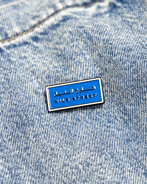 Blue enamel pin with nile street in text on a denim background inspired by sudanese culture on a brown bag as part of islamic relief uk new sudan souq collection
