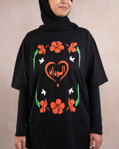 Woman wearing a black t-shirt with red hibiscus flower, white doves and heart design inspired by sudanese culture on a brown bag as part of islamic relief uk new sudan souq collection