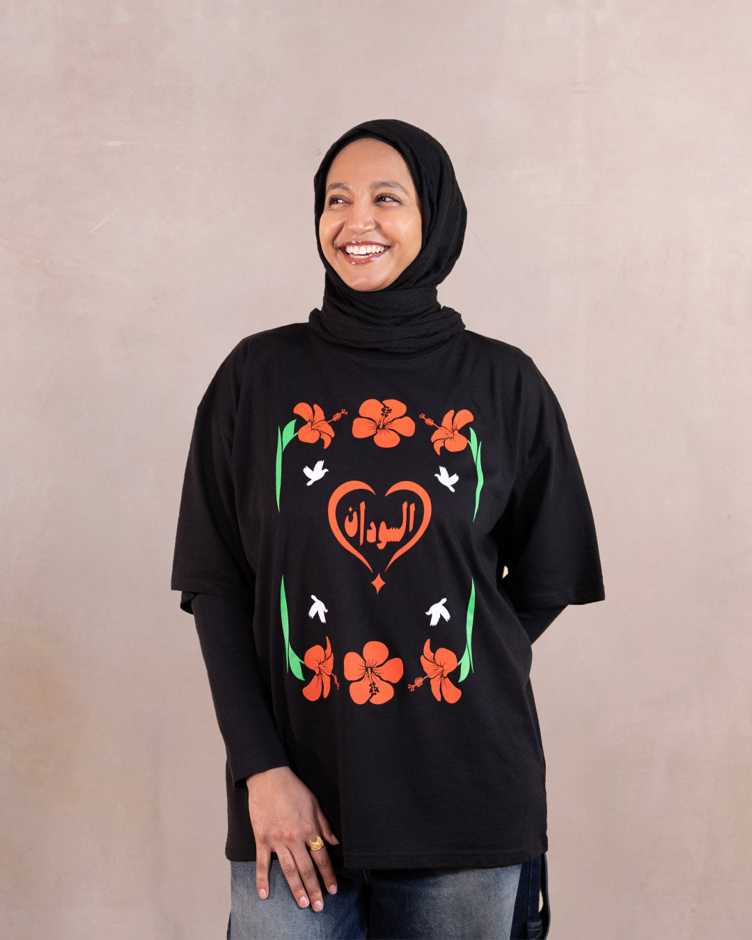 Woman wearing a black t-shirt with red hibiscus flower, white doves and heart design inspired by sudanese culture on a brown bag as part of islamic relief uk new sudan souq collection