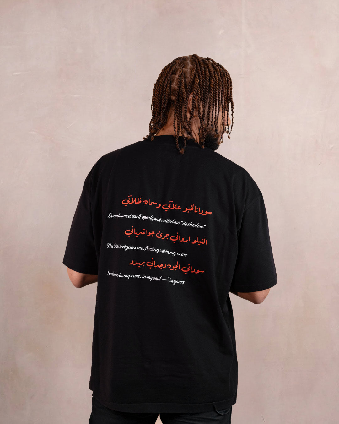 Back of man wearing a black t-shirt with red hibiscus flower, white doves and heart design inspired by sudanese culture on a brown bag as part of islamic relief uk new sudan souq collection