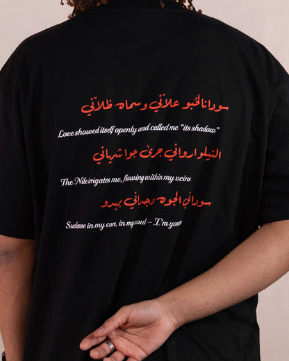 Close up of back text design of man wearing a black t-shirt with red hibiscus flower, white doves and heart design inspired by sudanese culture on a brown bag as part of islamic relief uk new sudan souq collection