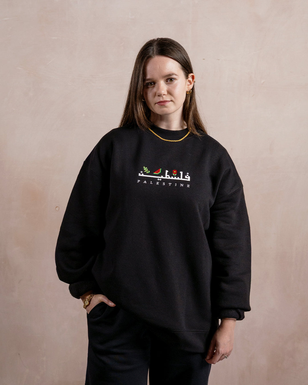 a woman wearing a black sweatshirt embroidered with the word palestine in english and arabic in white alongside an olive branch, red poppy and watermelon