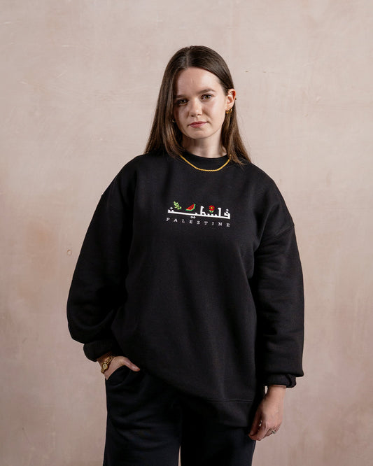 a woman wearing a black sweatshirt embroidered with the word palestine in english and arabic in white alongside an olive branch, red poppy and watermelon