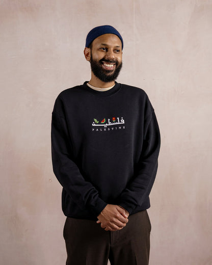 a man wearing a black sweatshirt embroidered with the word palestine in english and arabic in white alongside an olive branch, red poppy and watermelon
