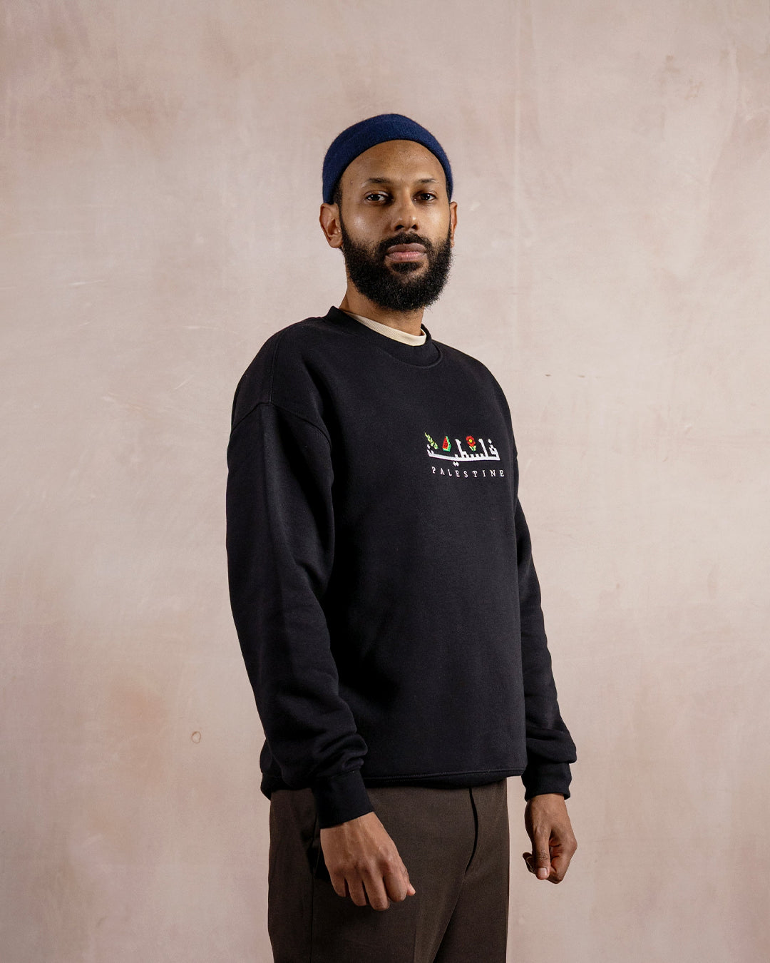 a man wearing a black sweatshirt embroidered with the word palestine in english and arabic in white alongside an olive branch, red poppy and watermelon