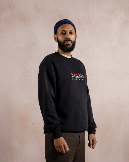 a man wearing a black sweatshirt embroidered with the word palestine in english and arabic in white alongside an olive branch, red poppy and watermelon