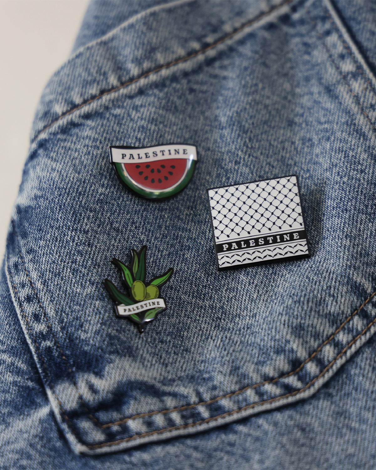 three different pin badges inspired by palestinian culture such as the watermelon, an olive branch and the keffiyeh print placed on a denim jacket