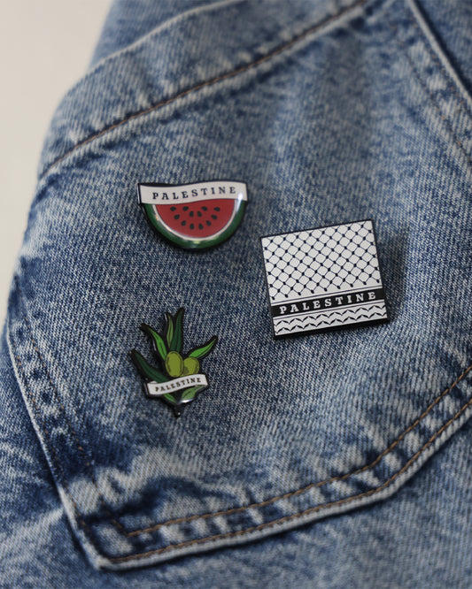 three different pin badges inspired by palestinian culture such as the watermelon, an olive branch and the keffiyeh print placed on a denim jacket