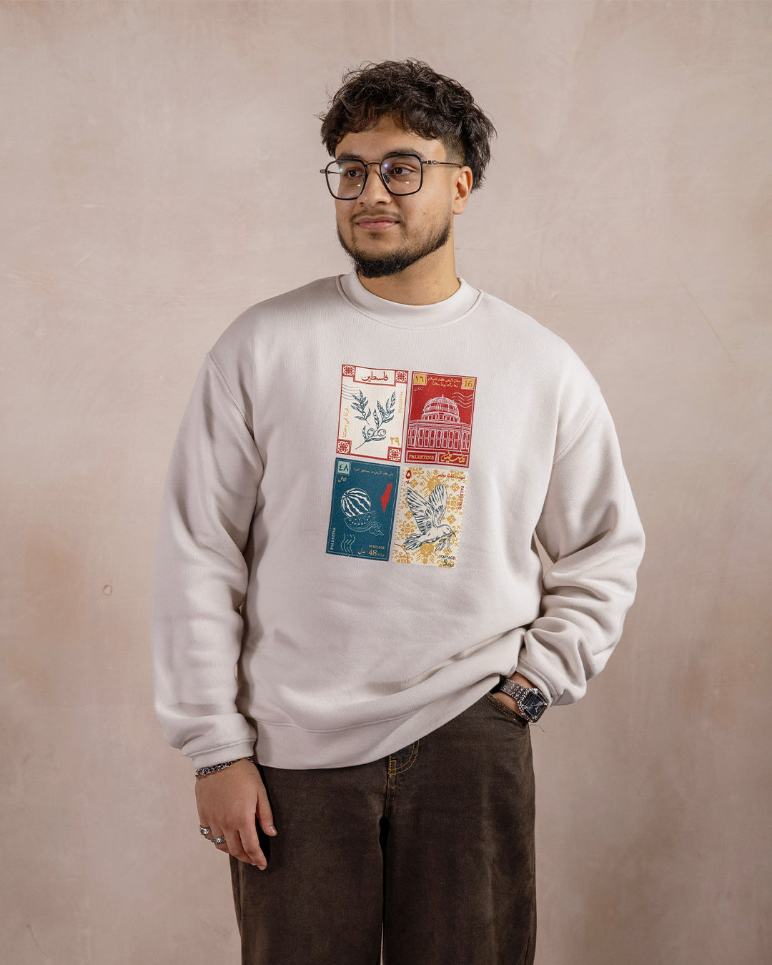a man wearing a light beige sweatshirt printed with a design inspired by palestinian culture with one hand in his pocket